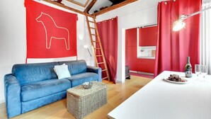 Una sala de estar moderna con un sofá azul, un cuadro abstracto rojo, una escalera de madera y una mesa blanca con una botella y vasos.