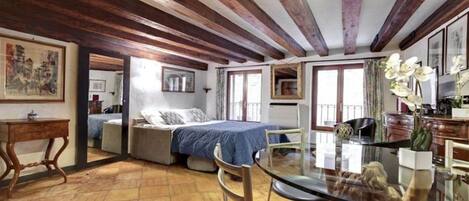 Una habitación con vigas de madera a la vista, una cama con cubrecama azul, una mesita de vidrio y una silla.