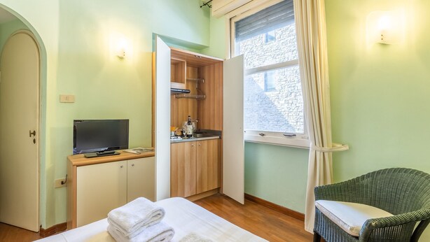 A room with a bed, a desk with a computer, a chair, a window with a view of a stone building, and a small kitchenette.