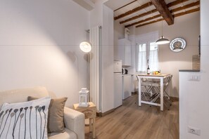 Un salon moderne avec un canapé, une petite table sur laquelle se trouvent une bouteille et des fruits, un coin cuisine équipé d’un réfrigérateur et d’un four à micro-ondes, ainsi qu’un luminaire suspendu.