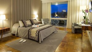 Une chambre d’hôtel avec un grand lit, un bureau, une chaise et une vue sur le paysage urbain illuminé la nuit.
