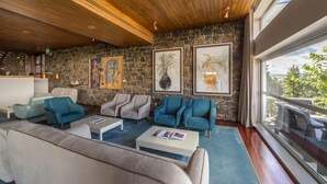 Un salon moderne aux murs en pierre, au plafond en bois et aux grandes fenêtres donnant sur un paysage verdoyant.