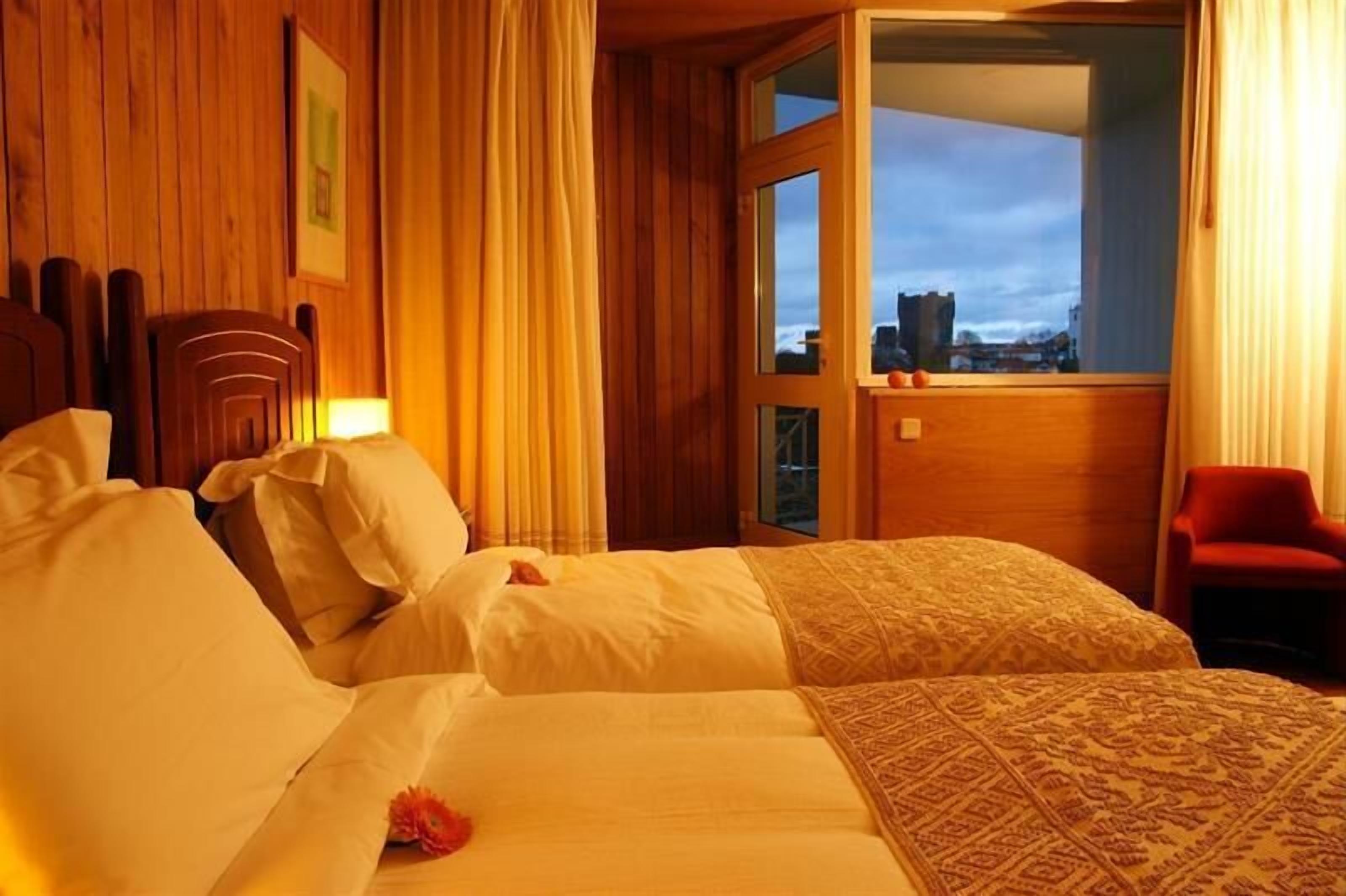 Ein Hotelzimmer mit Holzwänden, einem großen Bett mit weißer Bettwäsche, einem roten Sessel und Blick auf eine Stadt durch eine Glasdoor.