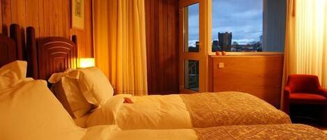 Ein Hotelzimmer mit Holzwänden, einem großen Bett mit weißer Bettwäsche, einem roten Sessel und Blick auf eine Stadt durch eine Glasdoor.