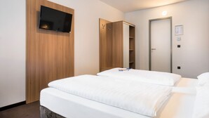 A hotel room with a bed, a TV mounted on a wooden panel, a wardrobe, and a door with a light. - Garner Hotel Stuttgart - Zuffenhausen By IHG (Stuttgart)
