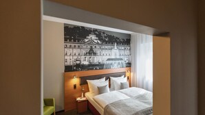 A hotel room with a bed, a nightstand, a lamp, a green chair, and a large cityscape mural on the wall. - Garner Hotel Stuttgart - Zuffenhausen By IHG (Stuttgart)