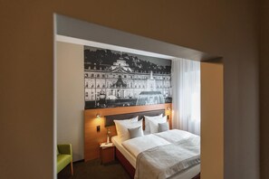 Ett hotellrum med en säng, ett nattduksbord, en lampa, en grön stol och en stor väggmålning som föreställer en stadsbild.