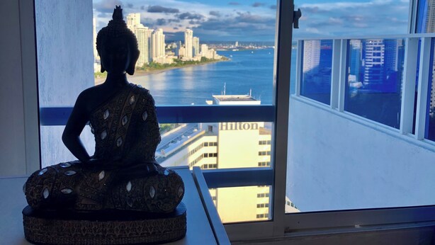 Eine Buddha-Statue auf einem Sockel neben einem Fenster mit Blick auf eine Stadtlandschaft und Wasser.