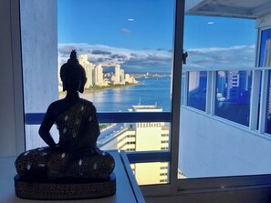 A Buddha statue on a pedestal by a window overlooking a cityscape and water. - SkyLaguito Ocean View 20 - Hilton District (Cartagea)