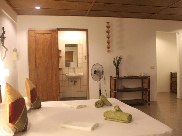 A hotel room with a bed, a wooden door, a bathroom with a sink and mirror, a fan, and a shelf with decorative items. - Chindonan Dive Resort (Coron)