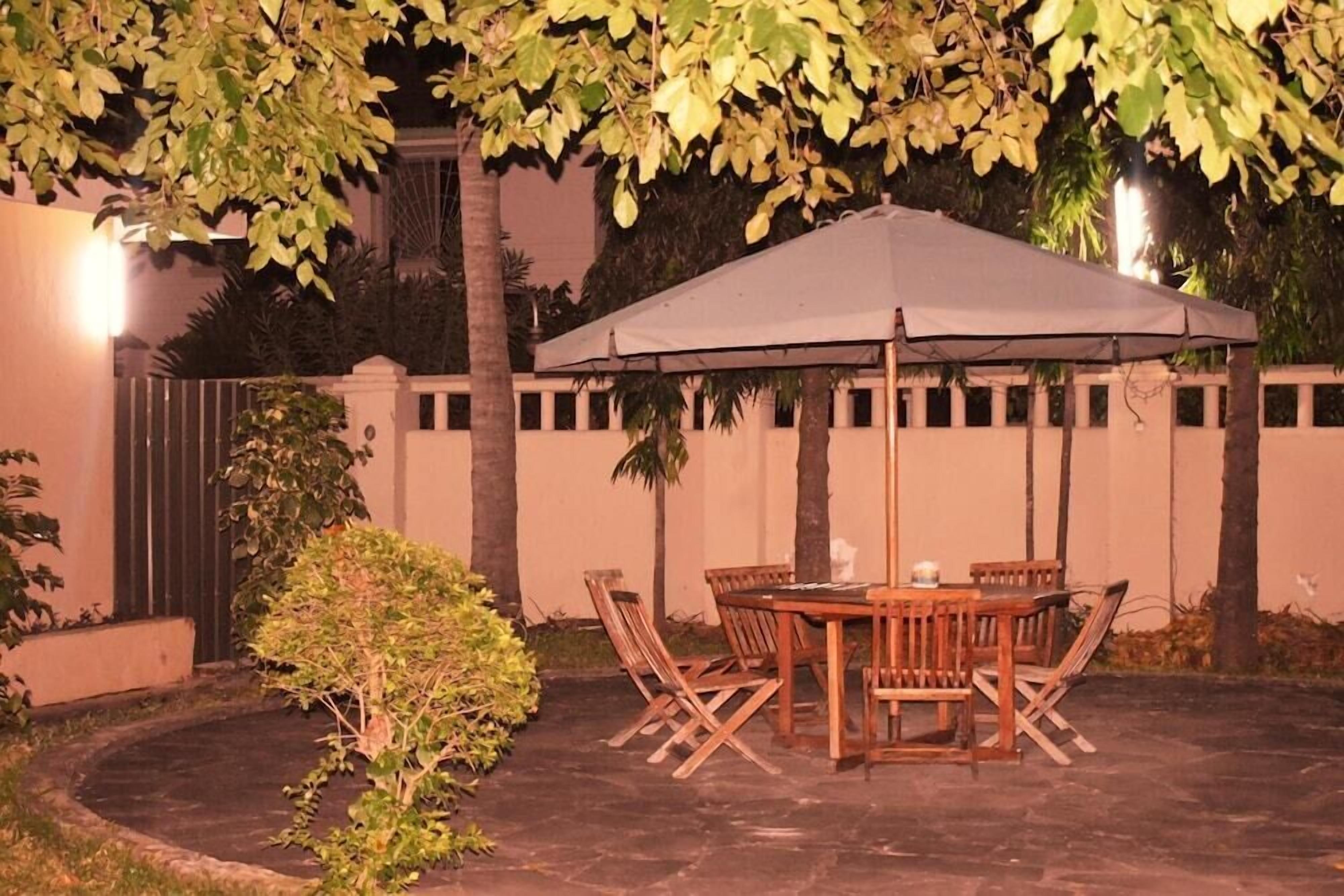 An outdoor patio with a wooden table and chairs under a large umbrella, surrounded by trees and a low wall.