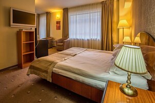 Una habitación de hotel con cama, escritorio, televisor, estantería y ventana con cortinas.