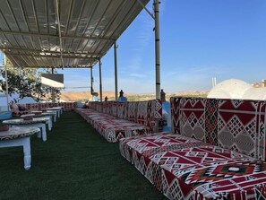 An outdoor seating area with patterned cushions and tables, under a shaded structure, with a view of distant buildings and hills. - Nubian Palace (Aswan)