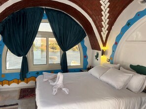 A bedroom with a large bed, a window with curtains, a view of a body of water, and a unique wall design. - Nubian Palace (Aswan)