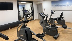 Ein Fitnessraum mit drei Ergometer, einem großen Spiegel und einem Fernseher an der Wand.