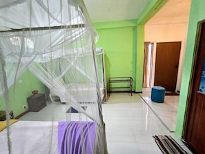 Una habitación con una cama bajo una mosquitera, un banco, una puerta y un cubo de basura azul.