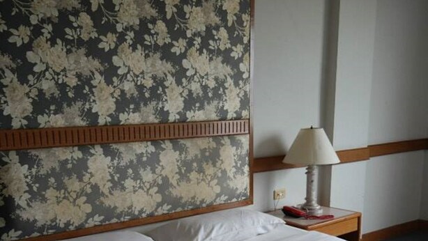 A neatly made bed with white linens, a wooden headboard with floral wallpaper, and a bedside table with a lamp and a red object.