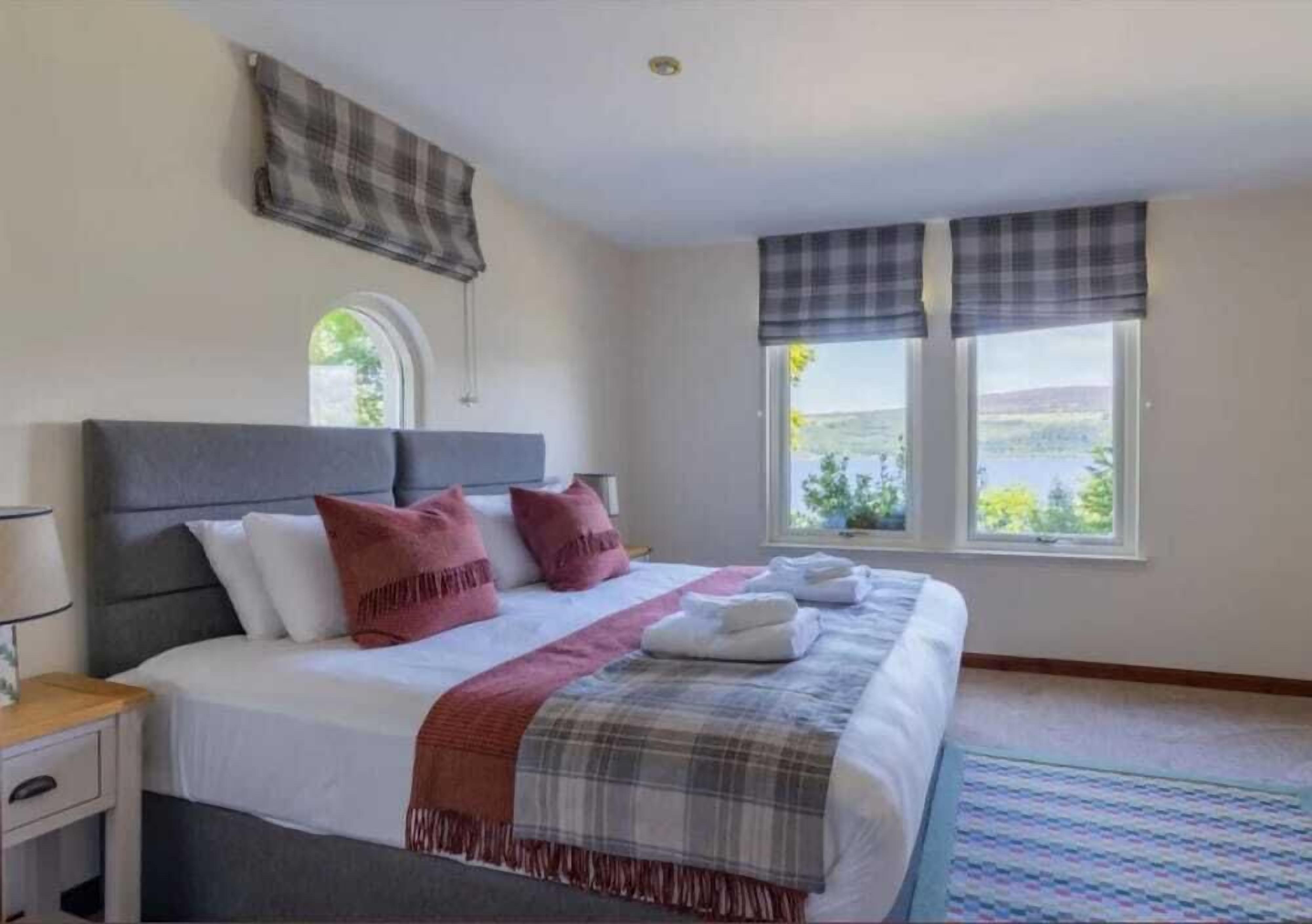 A neatly made bed with a grey headboard, white and red pillows, and a plaid blanket. There is a window with a view of greenery and a building.