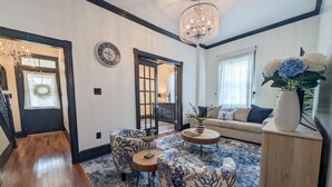 A living room with a chandelier, a sofa, patterned chairs, a coffee table, and a vase with flowers. - Updated 4BR/2BA Victorian - Linens Included, Beach Box, Walkable, Off-Street Parking (West Cape May)