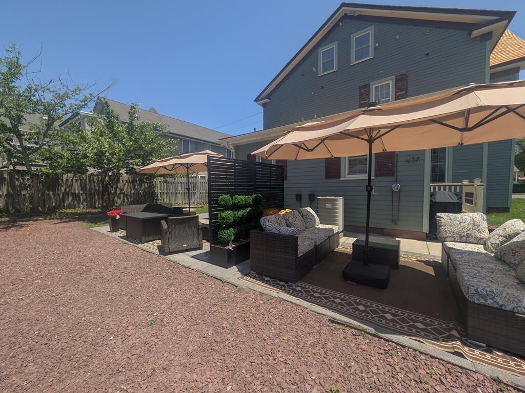 Une terrasse avec des meubles d’extérieur et des parasols, une clôture en bois et une maison portant le numéro 21.