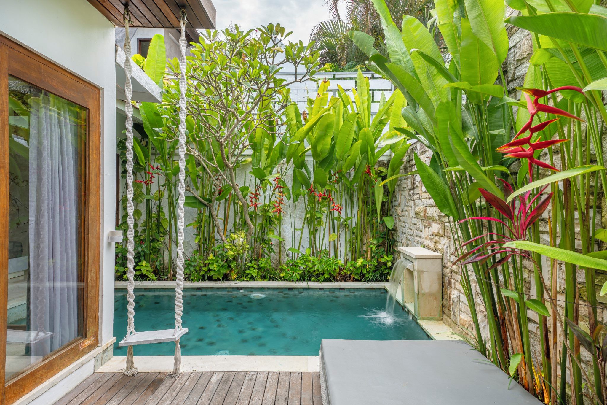 A pool area with a wooden deck, a swing, and lush tropical plants.