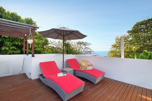 A balcony with two wicker lounge chairs, one with a red cushion and the other with a grey cushion, and a large umbrella providing shade. - The Jimbaran Beach Villa by Ini Vie Hospitality (Jimbaran)