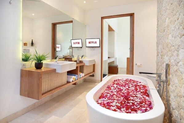 Un baño moderno con dos lavamanos, un espejo grande, una bañera llena de pétalos de rosa y dos televisores empotrados que muestran 'Netflix'.