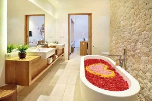 Une salle de bain moderne avec une baignoire remplie de pétales de rose, un meuble-lavabo en bois et un mur en pierre.