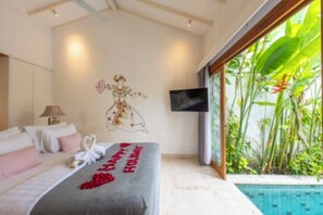 A hotel room with a bed, a wall-mounted TV, and a view of a swimming pool. - The Jimbaran Beach Villa by Ini Vie Hospitality (Jimbaran)