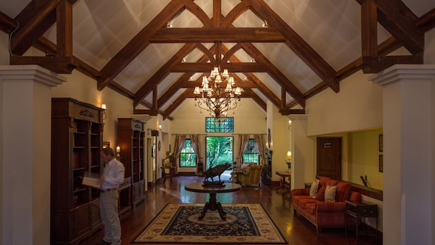 A spacious lobby with high ceilings, exposed wooden beams, a chandelier, a large rug, a sofa, and a fireplace. - The Manor Ngorongoro (Karatu)