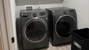 A laundry room with two front-loading washing machines, a white cabinet above, and a black laundry basket. - Spacious 4Bd/4bath Home with Media Room-Great for Master’s Week! (Evans)