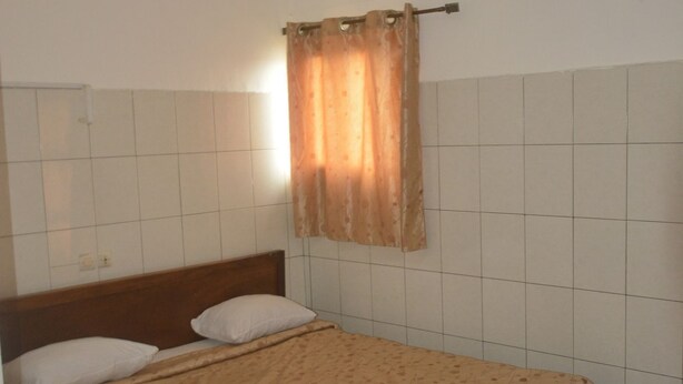 A single bed with a wooden headboard, a beige bedspread, and a white pillow. There is a window with a brown curtain, and an air conditioning unit mounted on the wall. - Hotel La Citadelle (Abidjan)