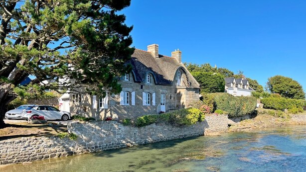 Een stenen huis met vrij uitzicht op de rivier, een geparkeerde auto en een grote boom op de voorgrond.