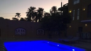 A swimming pool with blue lighting at dusk, surrounded by palm trees and a building with Egyptian-themed wall art.