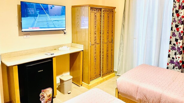 A hotel room with a television on the wall, a wooden wardrobe, a small refrigerator, a waste bin, and two beds with pink bedding.