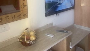 A hotel room with a television mounted on the wall displaying a show, a wooden headboard with carvings, a small table with a basket of fruits, and a trash bin.