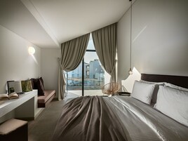 Quarto de hotel moderno com uma cama grande, uma escrivaninha com um laptop, uma cadeira e vista para a paisagem urbana através de grandes janelas.