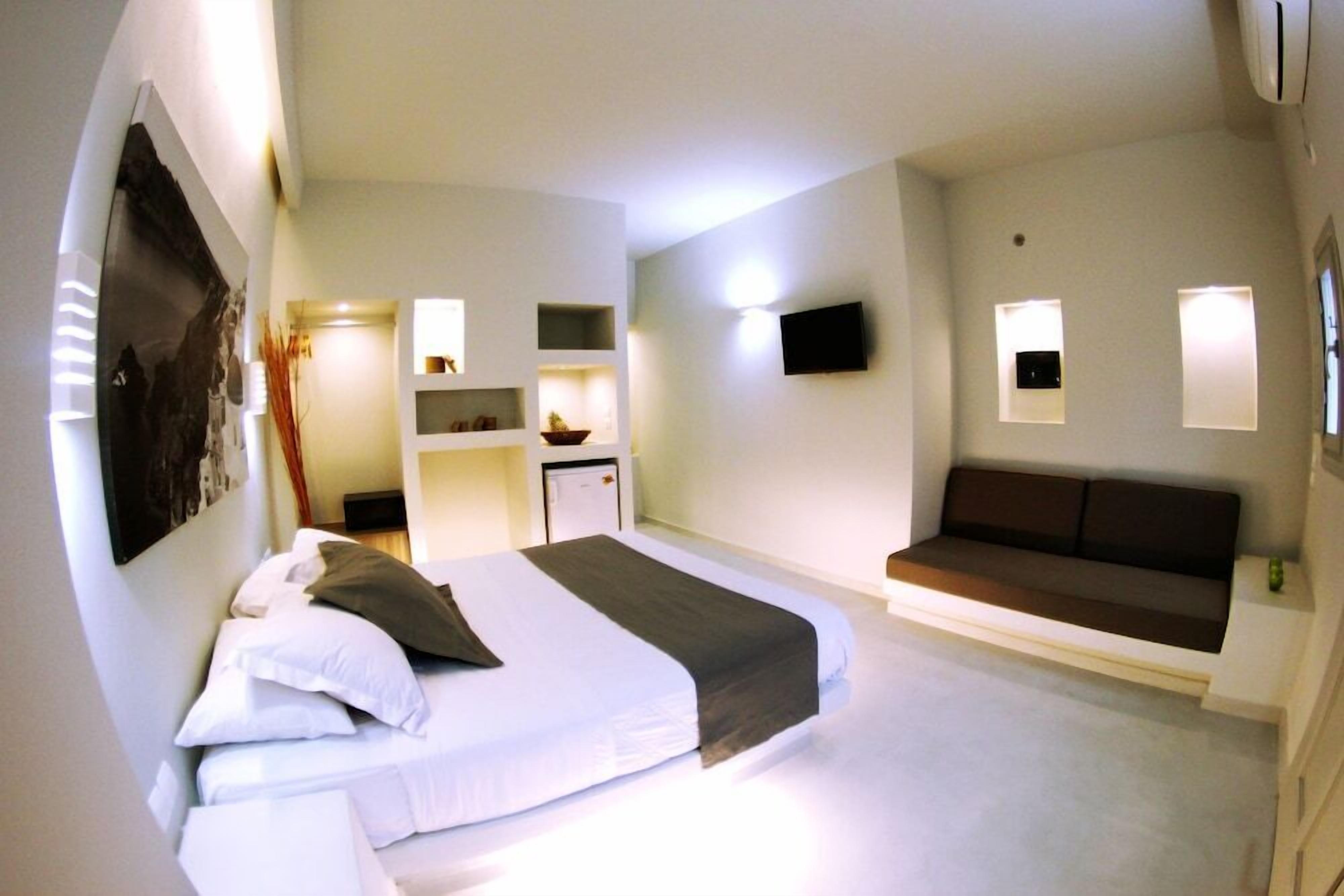 A modern hotel room with a large bed, a sofa, a desk with a chair, and a flat-screen TV mounted on the wall.