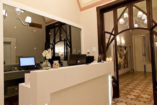 A hotel reception area with a white counter, a computer, a vase with flowers, and a large mirror. - Palazzo Arco Cadura Hotel & Spa (Galatina)