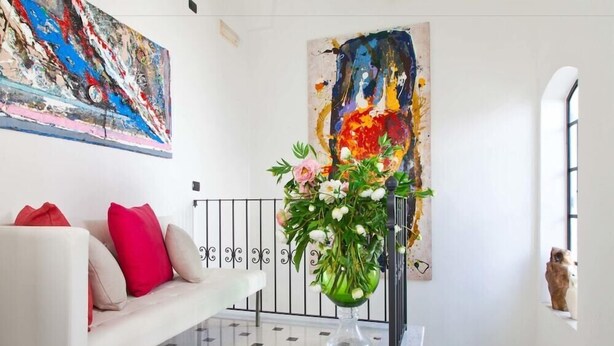 Een moderne woonkamer met een witte bank, rode kussens, een glazen vaas met bloemen en abstracte schilderijen aan de wanden.