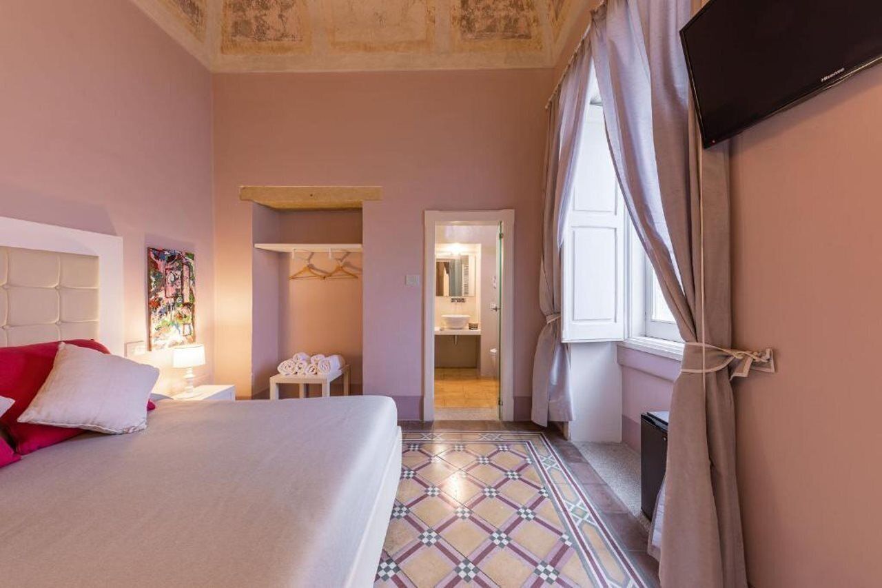 Habitación de hotel con cama, mesita de noche, lámpara, perchero y baño visible a través de una puerta abierta.