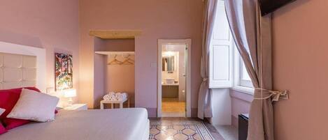A hotel room with a bed, a nightstand, a lamp, a hanging coat rack, and a bathroom visible through an open door.