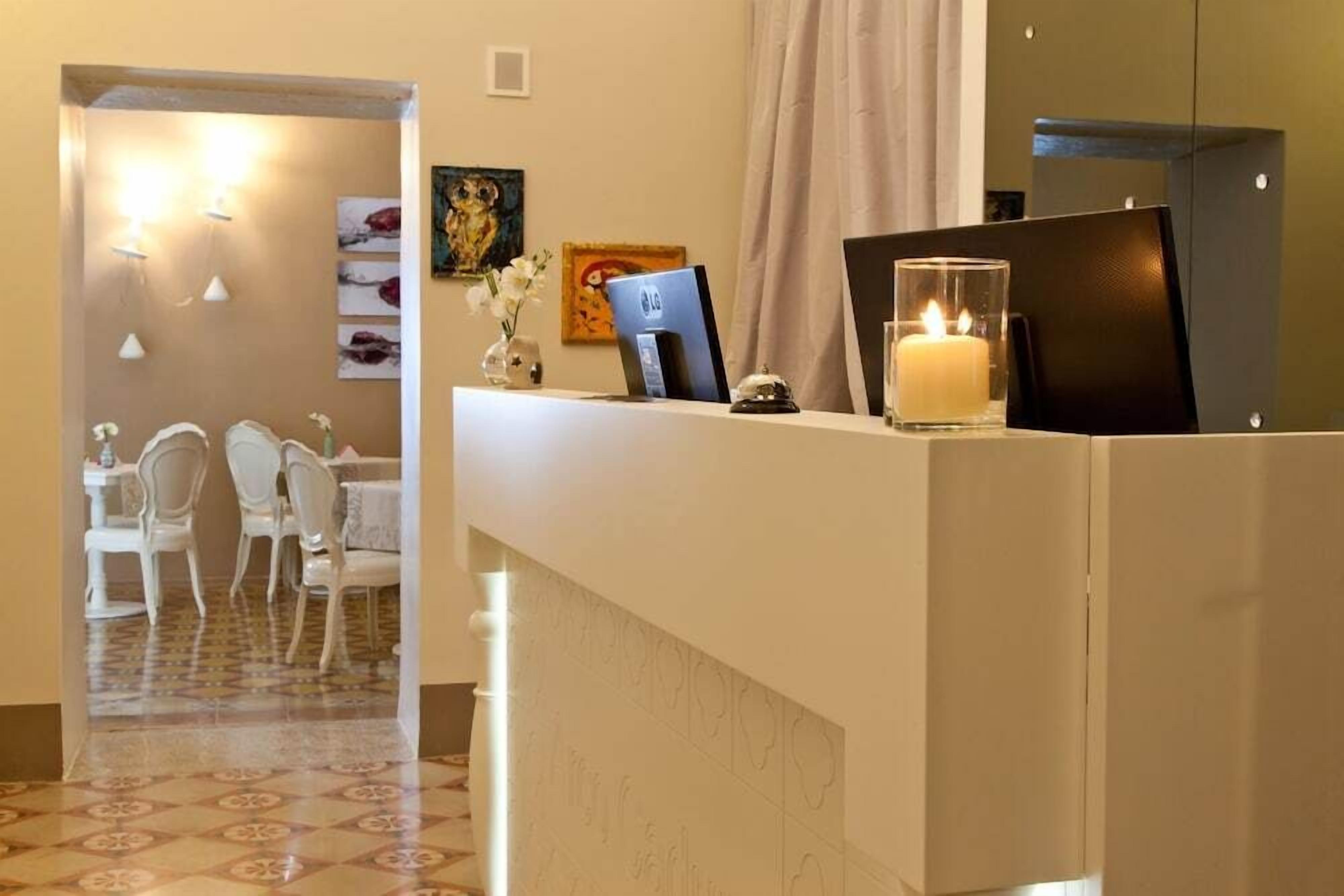A hotel reception area with a reception desk, a computer, a candle, and a vase with flowers. There is a dining area with white chairs and a table in the background.