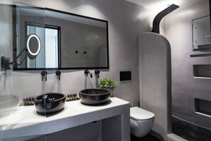 Ein modernes Badezimmer mit einem großen Spiegel, zwei schwarzen Schalen auf der Ablage, einer Toilette und einer Dusche mit einer gewölbten Wand.