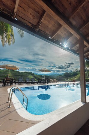 Une piscine extérieure dotée de chaises longues et de parasols, entourée d’une terrasse en bois et de palmiers.