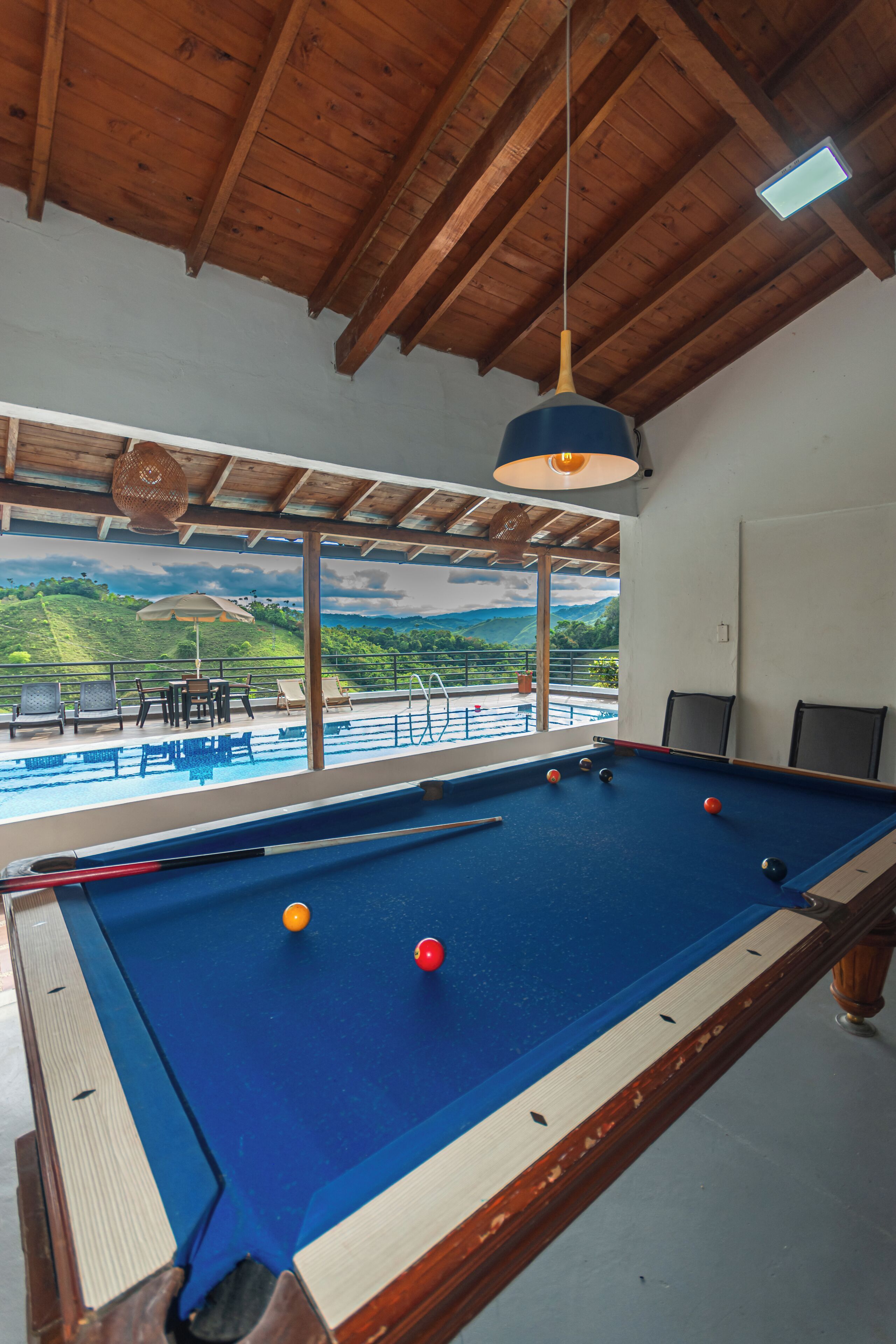 Una mesa de billar con bolas, una lámpara colgante y vista a un área de piscina con sillas y una mesa.