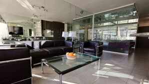 Ein modernes Wohnzimmer mit Glastisch, Ledersofa und einem durch Glaswände sichtbaren Küchenbereich.