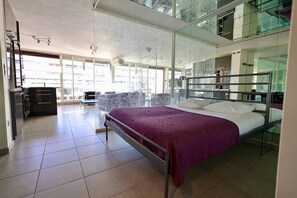Habitació d'hotel moderna amb un llit gran, una taula de treball i una cadira. L'espai disposa d'ampies finestres i un balcó.