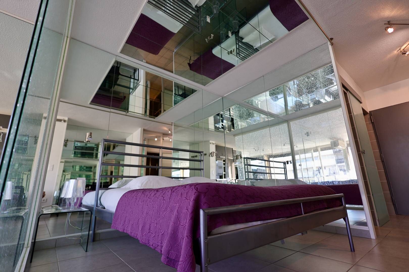 A modern hotel room with a large bed, a glass partition, and a view of the outdoors.