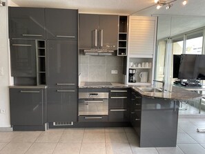 A modern kitchen with dark cabinetry, a central island, and a built-in oven. - RIFLESSO - Héliopolis D 70 - Luxe - 2 people (AGDE)